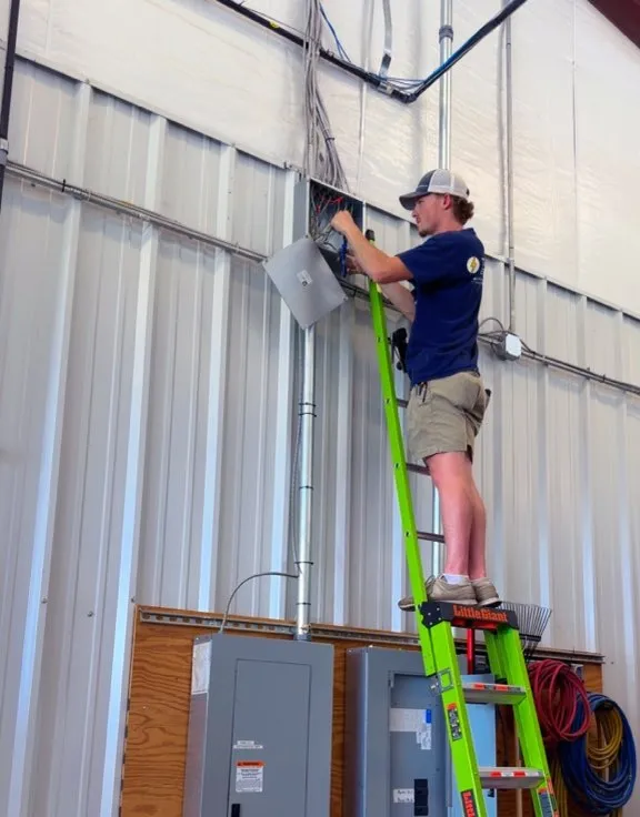 A worker on a green ladder at an open electrical enclosure with metal conduit running along a metal building wall
