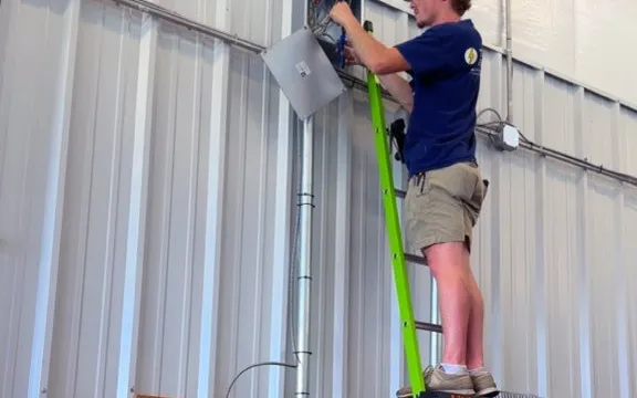 A worker on a ladder beside conduit and an open electrical box.
