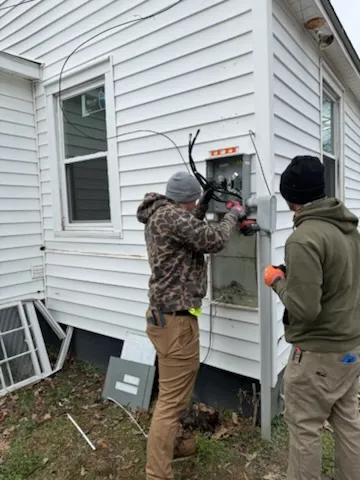 Two workers attending to an open electrical service panel on the side of a white-sided house