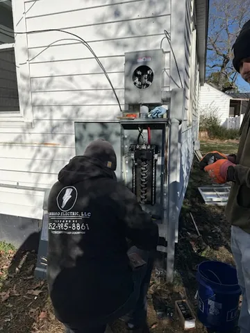 Two workers beside an open electrical panel and meter enclosure on a white-sided house