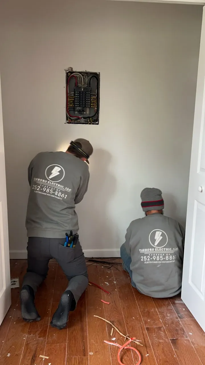 Two workers kneeling on a wood floor below an open electrical panel set into a painted interior wall