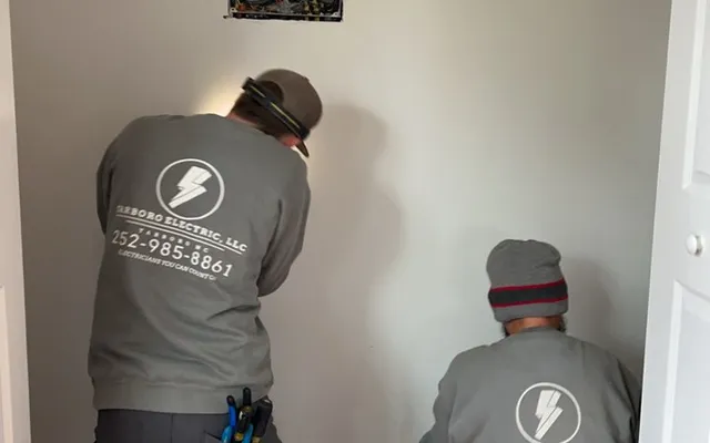 Two workers working below an open electrical panel during an indoor renovation.