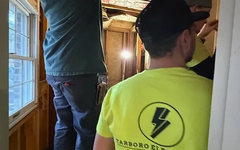 Workers in an unfinished room with visible wiring and exposed timber framing.