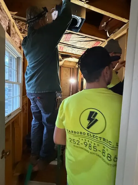 Workers in an unfinished room with exposed wood framing, ceiling wiring and insulation
