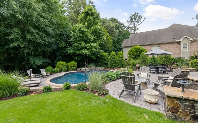 A backyard swimming pool surrounded by a stone patio, chairs and garden planting.